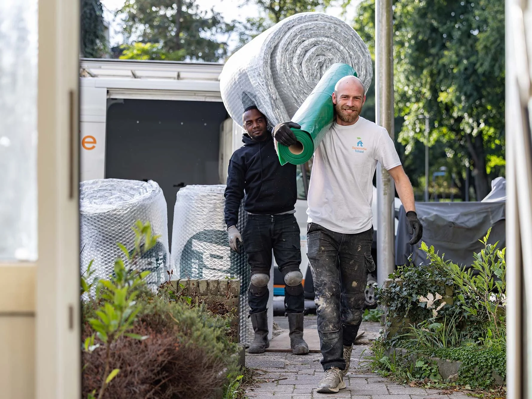 Twee medewerkers van Renovatie Totaal dragen rollen isolatiefolie van de bus naar een woning