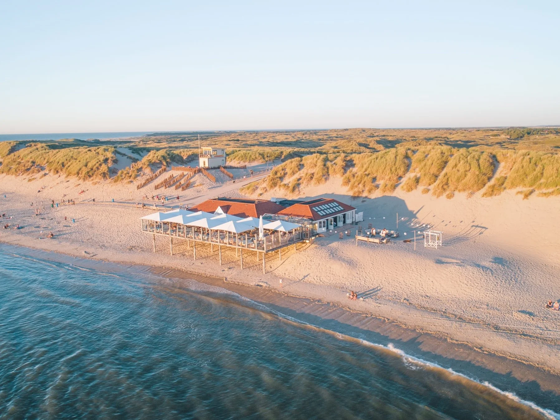 Luchtfoto van een strandpaviljoen tussen de duinen van Ameland, met de rood-witte vuurtoren op de achtergrond